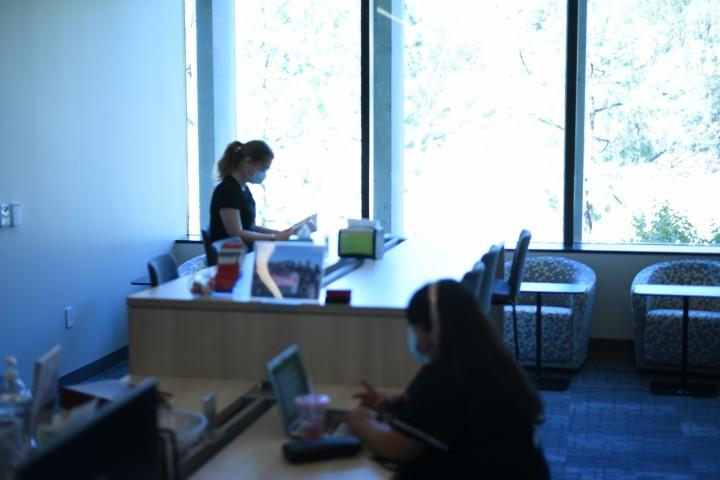 Student with headphones sits at table reading. Student in the background sits at tall table reading. 